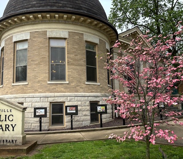 A picture of the northeast corner of the library building, the library sign, and a flowering pink dogwood tree.